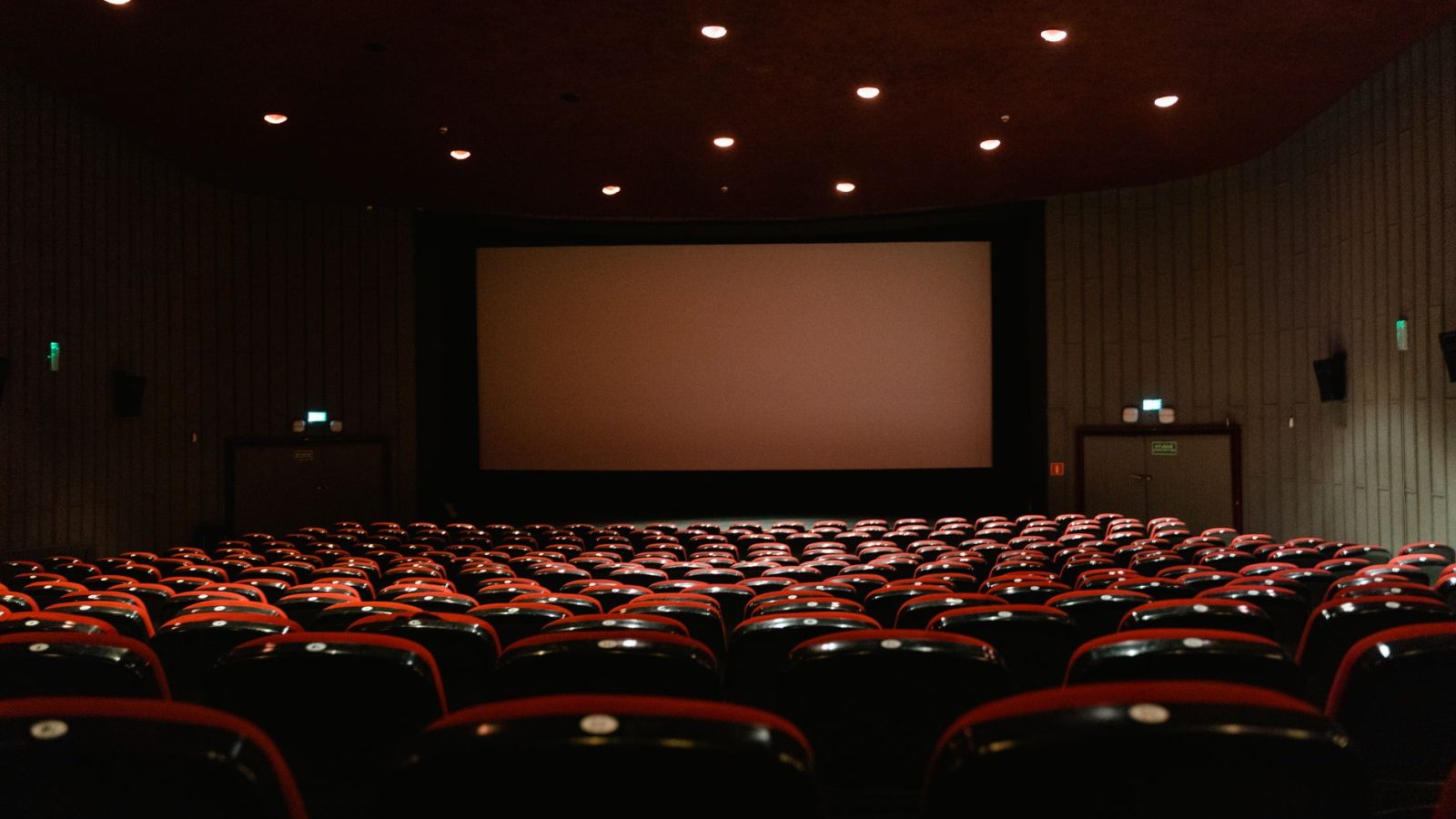 A dimly lit empty cinema theater with rows of red chairs facing a projector screen.