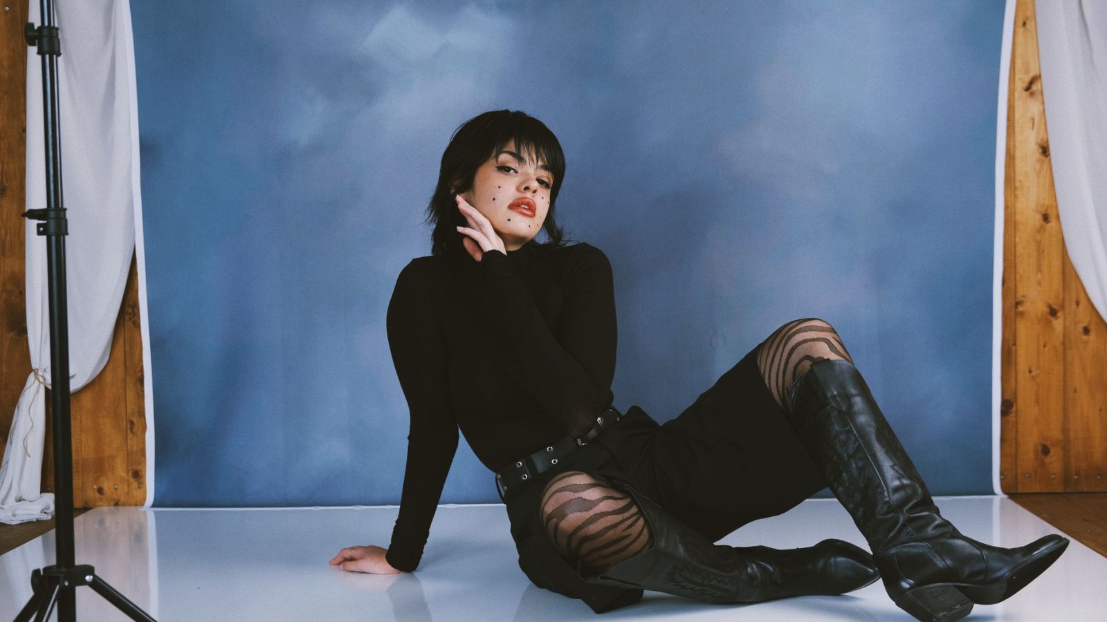 A fashionable woman posing indoors against a blue backdrop wearing black boots and stylish clothing.