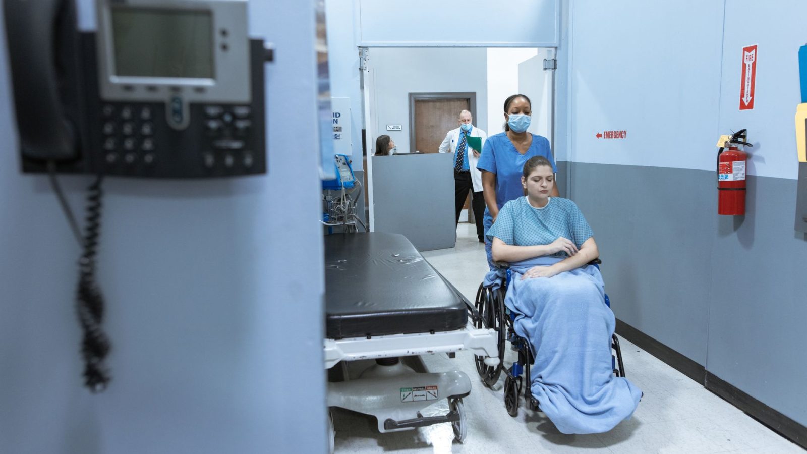 A nurse helps a patient in a wheelchair down a hospital corridor, reflecting care and medical professionalism.