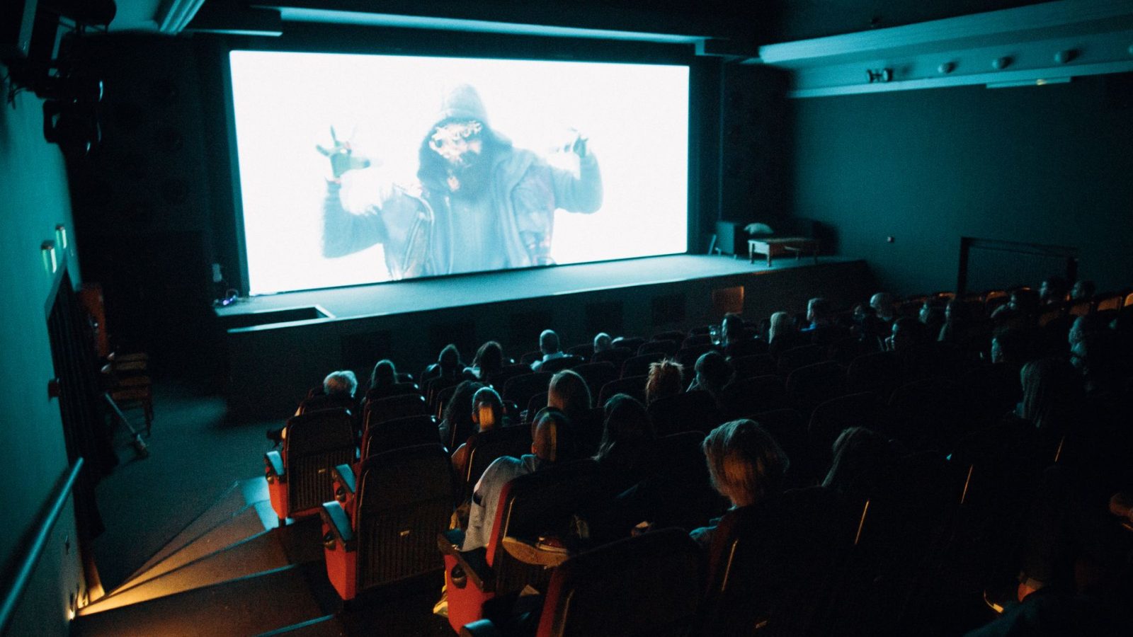 Crowded cinema theater with audience watching a movie on a large screen in a dark setting.