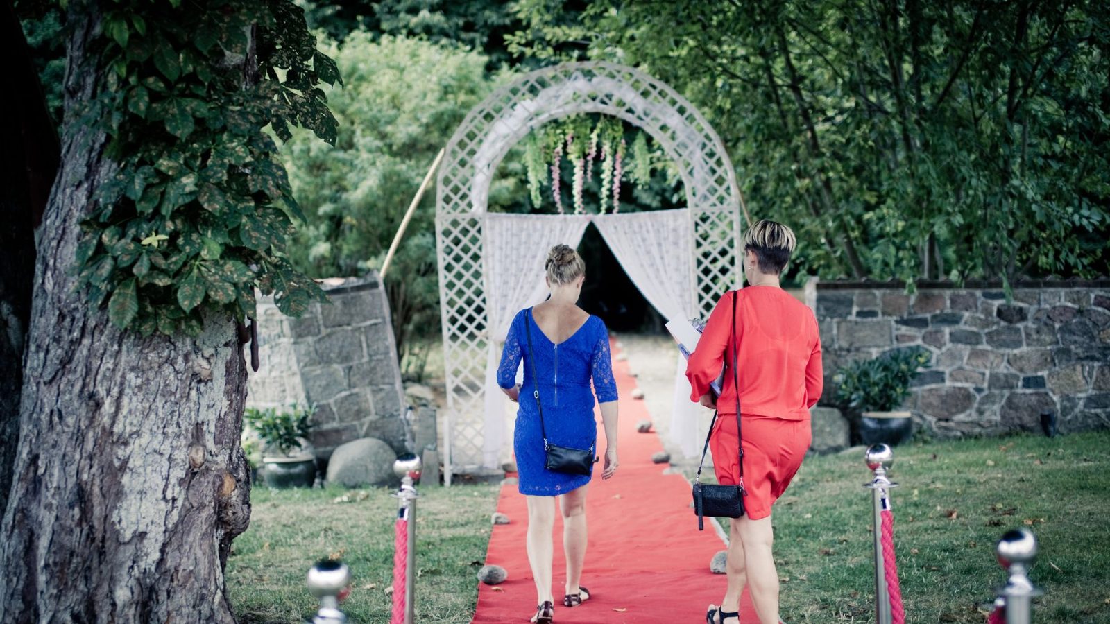 Two women walk on a red carpet through a garden towards a decorative archway.