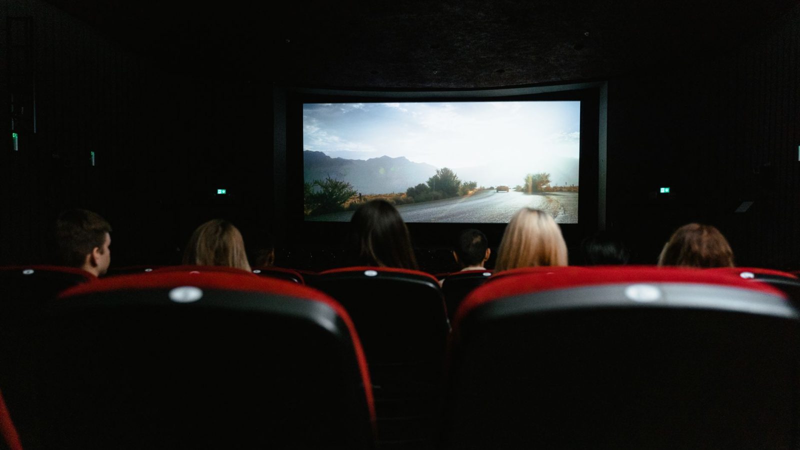 View from behind of people watching a movie in a cinema with red seats and a large screen.