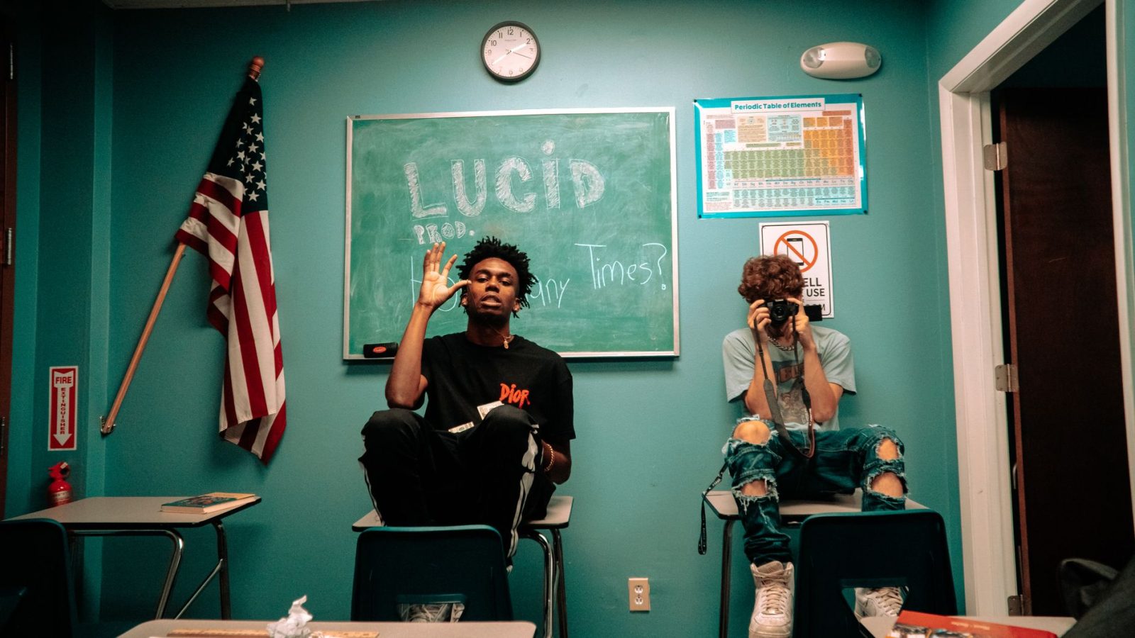 Two teenagers in a classroom setting, one waving and one photographing, with USA flag.